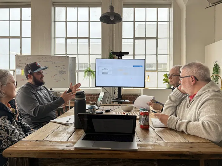 Team members discuss their work around a table with laptops and a shared screen.