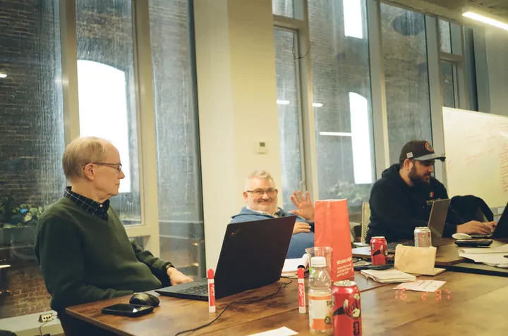 Three team members work at a shared table, with one waving toward the camera.